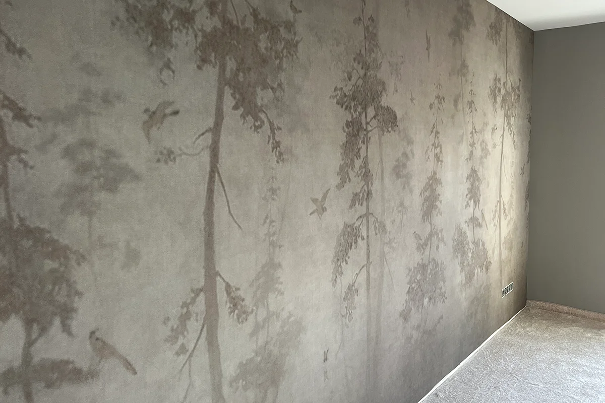 Eine Wand mit einer dezenten, gedämpften Tapete. Die Tapezierarbeit von der Maler- und Glaserwerkstatt Förster aus Nideggen zeigt Silhouetten von Bäumen und Vögeln und schafft so ein sanftes, von der Natur inspiriertes Muster in einem neutral getönten Raum mit Teppichboden.