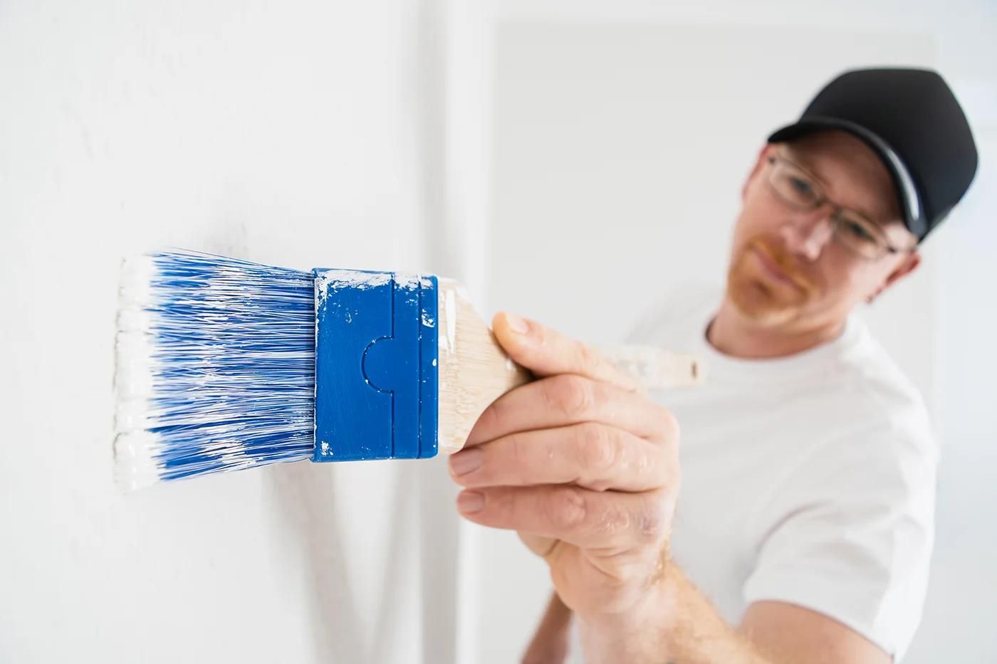 Ein Mitarbeiter des Meisterbetriebes Förster aus Nideggen mit Brille, schwarzer Mütze und weißem Hemd bemalt mit einem großen blauen Pinsel eine weiße Wand und arbeitet konzentriert an dem Innenanstrich in Düren.