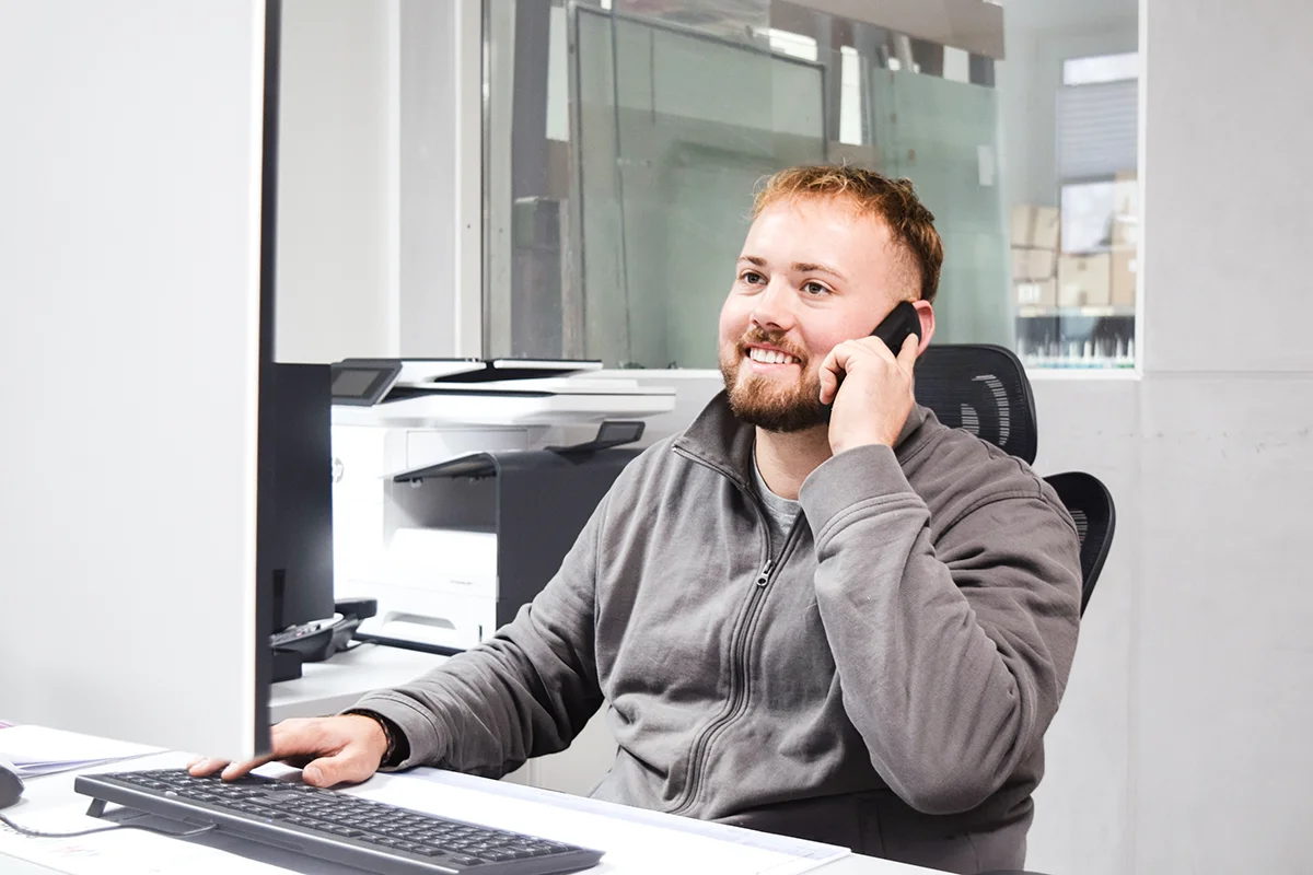 Ein bärtiger Mann in einer grauen Jacke mit Reißverschluss sitzt in einem modernen Büro einer Maler- und Glaserwerkstatt in Nideggen an einem Schreibtisch, lächelt, telefoniert und berät einen Kunden zum Thema Fassadenanstrich.